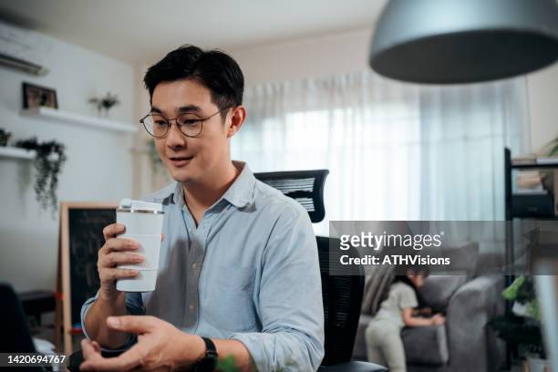 man explaining business plan to his colleagues on video conference business meeting via laptop computer while his daughter using phone on sofa in the living room at home. - young businessman using a virtual screen stockfoto's en -beelden