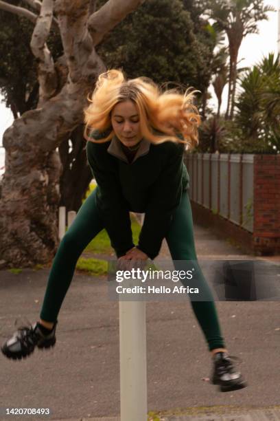 front view of a young woman in the process of leap-frogging an upright white pole with blond untied hair raised up in the air, a teenage girl dressed in green having fun on a suburban sidewalk with cars seen parked in the street - vaulting gymnastics stock pictures, royalty-free photos & images