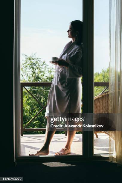 young woman in white bathrobe in hotel holding cup with morning coffee - breakfast with view stock-fotos und bilder