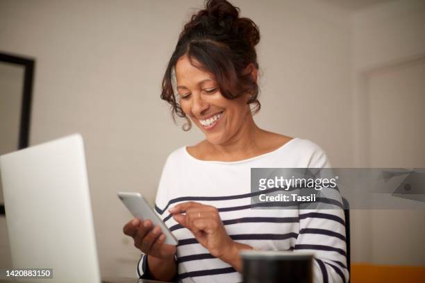 mature woman texting on a phone while relaxing at home in a living room, smiling and looking happy. cheerful female enjoying an easy online dating app conversation, satisfied with streaming service - dating stockfoto's en -beelden