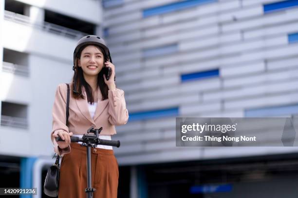 asian smiling young woman with helmet hold mobile smart phone talk with business during go to office work at street with electric  scooter, eco friendly - オーバーコート ストックフォトと画像