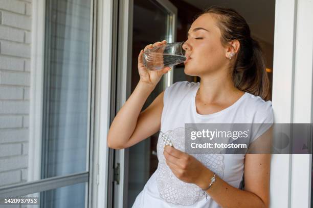 young woman standing with eyes closed, drinking water after taking a pill for her headache - birth control pill stock pictures, royalty-free photos & images