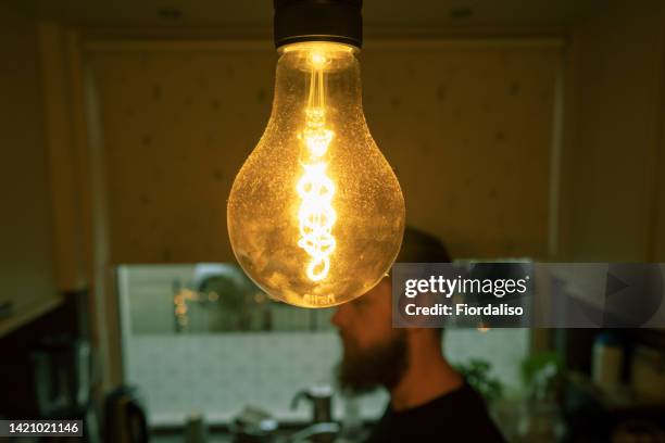 a bearded man with long curly hair in a bun standing by the window behind a large yellow light bulb with a spiral wick. the concept of saving electricity, growing rent bills, economic crisis, unemployment - electricity bill stock pictures, royalty-free photos & images