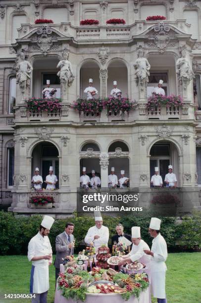 Brigade de cuisine, whisking and holding kitchen utensils, look out of the windows of the Brenner's Park Hotel, as chefs set up a buffet table in the...