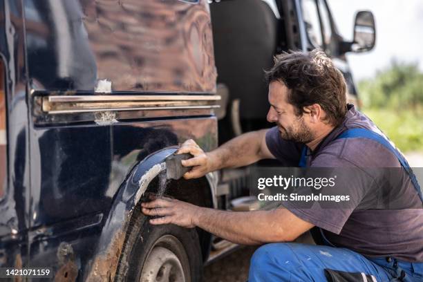 car mechanic scrubs rust off the fender of an old black van outdoors. - rusty old car fotografías e imágenes de stock
