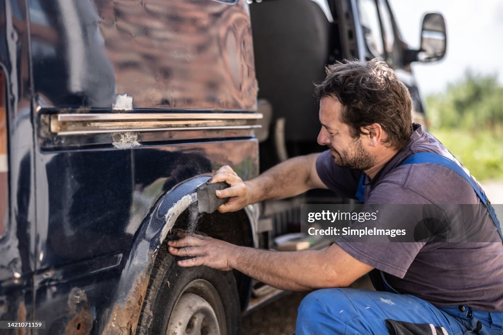 Car mechanic scrubs rust off the fender of an old black van outdoors.