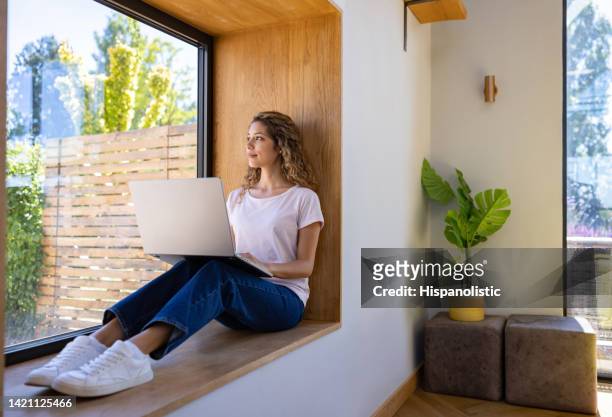 thoughtful woman at home working on her laptop by the window - description bildbanksfoton och bilder