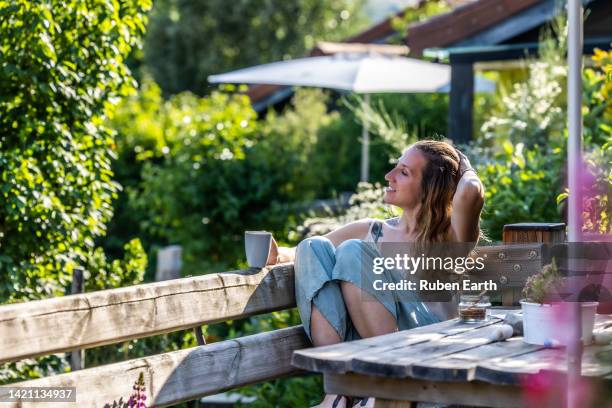 young woman relaxing with a cup of coffee while sitting on a wooden bench in a garden - lentezza foto e immagini stock