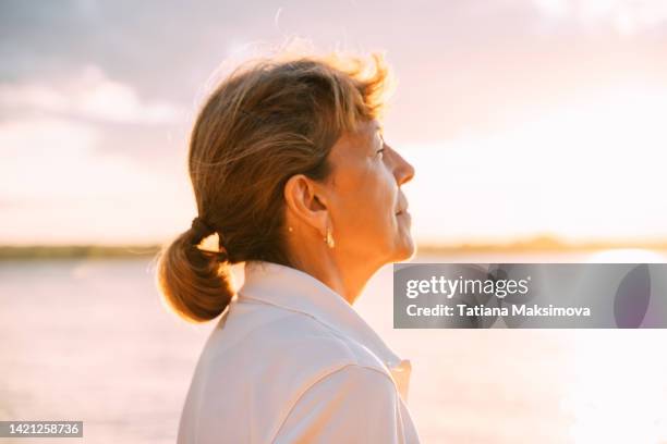 senior woman enjoys a moment at sunset on the river beach. close-up portrait. copy space. - mature woman - fotografias e filmes do acervo