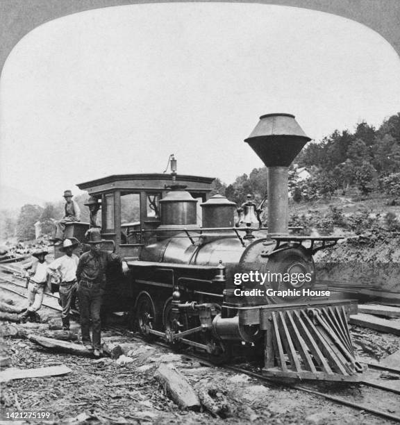 Men pose beside an steam locomotive with a large spark-arresting chimney and a cowcatcher mounted to its front, designed to deflect obstacles on the...