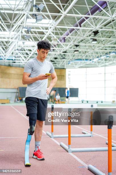 young fitness man with physical disabilities and prosthesis using cell phone at hurdle training - amputé photos et images de collection