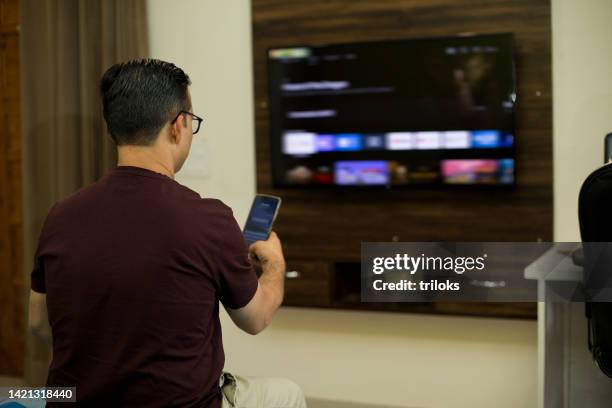 mann, der ein mobiltelefon benutzt, während er zu hause im wohnzimmer sitzt - indian living room stock-fotos und bilder