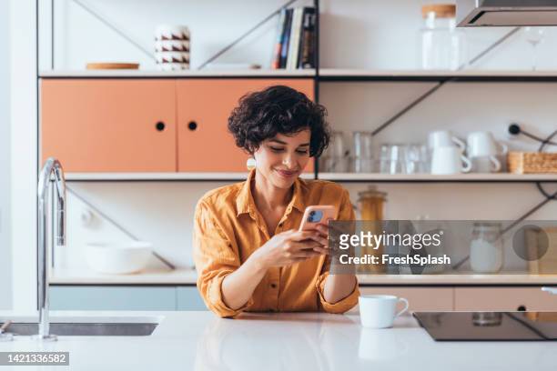a happy beautiful businesswoman sitting and enjoying drinking her first morning  coffee at home while texting on her mobile phone - leisure work coffee happy stockfoto's en -beelden