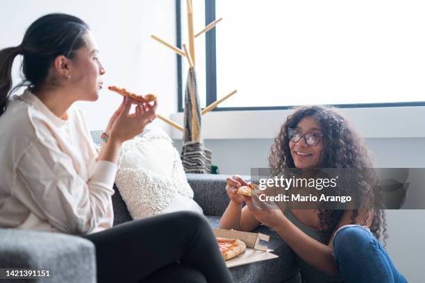 two latin friends spend time together in the living room of their house eating a piece of pizza - afro friends home stock pictures, royalty-free photos & images