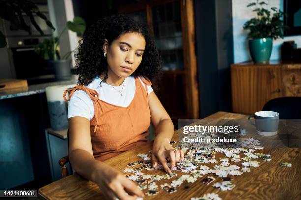 woman working on a puzzle at a table in the living room of her modern comfortable house. girl doing jigsaw game challenge for mental health and to relax while having cup of coffee in a lounge at home - puzzel stockfoto's en -beelden
