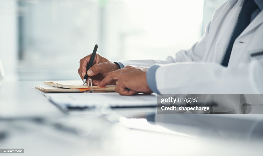 Doctor hands writing on paper or document at a desk in the hospital. Healthcare professional drafting a medical insurance letter, legal paperwork or form. A GP filing a document in a clinic office