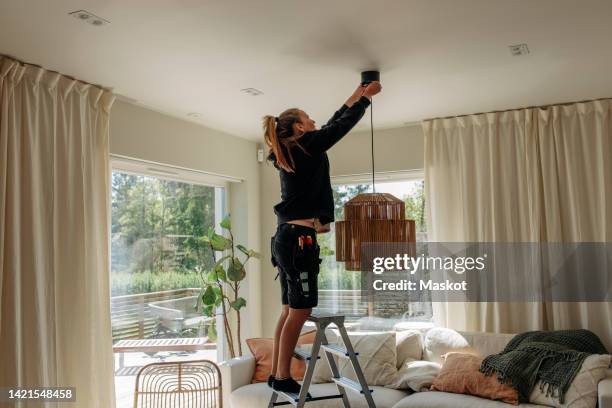 full length of female electrician installing pendant light while standing on ladder in living room at home - pendant light stockfoto's en -beelden
