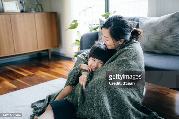 loving young asian mother and cute little daughter wrapped in blanket, relaxing at home, sitting on the floor in the living room. bonding between mother and daughter. family lifestyle. love and care concept - angelica hale fotografías e imágenes de stock