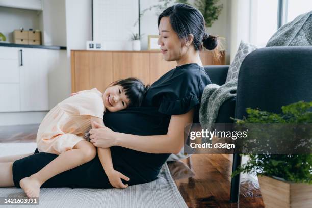 lovely little asian girl embracing her mother's pregnant belly, listening the heartbeat of her unborn sibling at home, looking at camera with smile. sibling love. welcoming a new family member with love and care concept - smile woman child stock-fotos und bilder