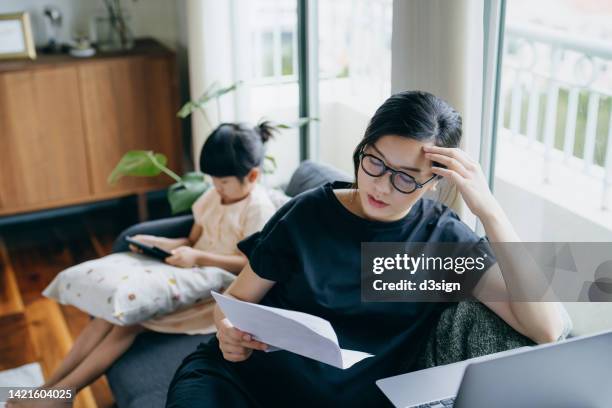 young asian mother looking stressed while managing her financial bills and tax documents, working from home on laptop and her daughter is using digital tablet in the background. working mother managing work life and childcare at home - stressed parent stock pictures, royalty-free photos & images