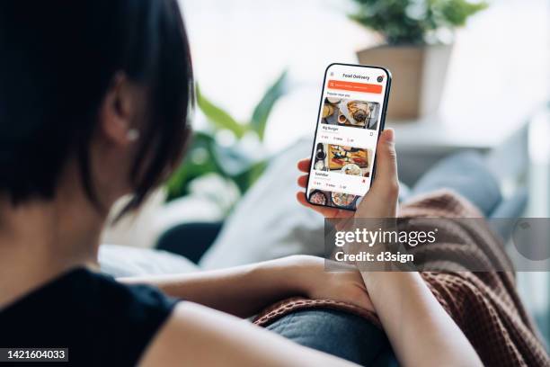 over the shoulder view of young asian woman sitting on the sofa, using meal delivery service and ordering food online with smartphone mobile app at home - mobile screen stockfoto's en -beelden