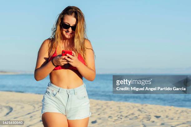 young woman walking with mobile phone on the beach - swimsuit models girls stock pictures, royalty-free photos & images