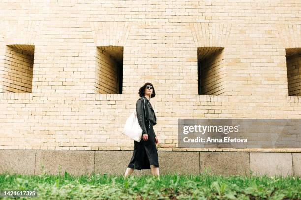 beautiful extravagant young woman is walking on brick wall background. she is wearing sunglasses, black dress, leather jacket and holding shopping bag on the shoulder. concept of fashionable youth style - leather dress stockfoto's en -beelden