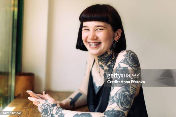 laughing beautiful extraordinary woman with many tattoos and short dark hair is sitting at a wooden table. she is wearing a black dress. concept of appearance that goes out of the frames - dress short sleeve stockfoto's en -beelden