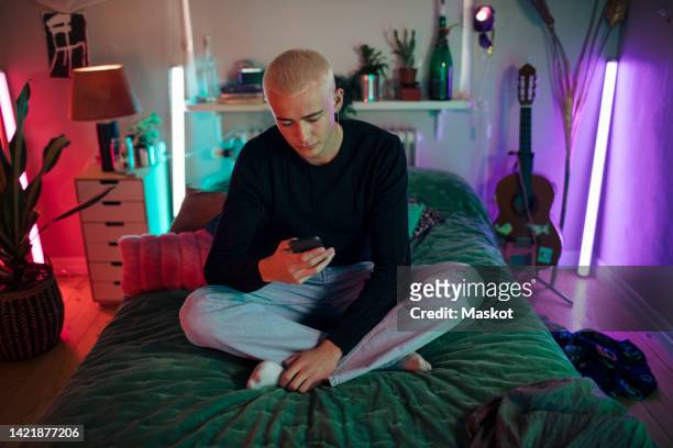 full length of young man using smart phone while sitting cross-legged on bed at home - generacion z fotografías e imágenes de stock