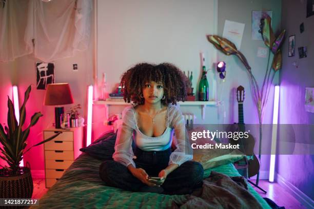 portrait of afro woman sitting with cross-legged on bed in illuminated bedroom at home - decolleté stockfoto's en -beelden