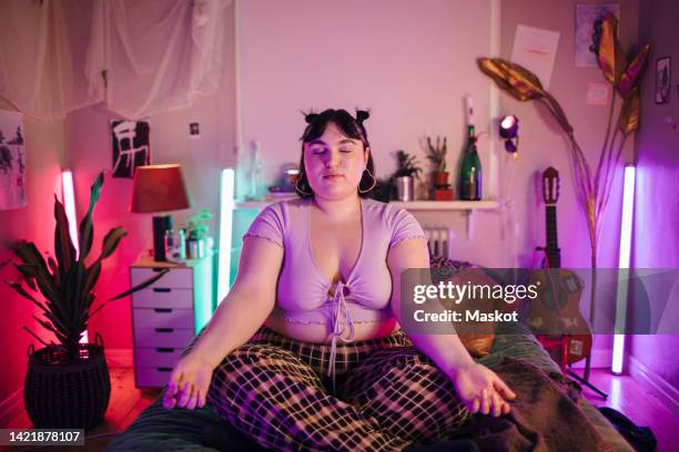 young woman meditating on bed in illuminated bedroom at home - mindfulness stock-fotos und bilder