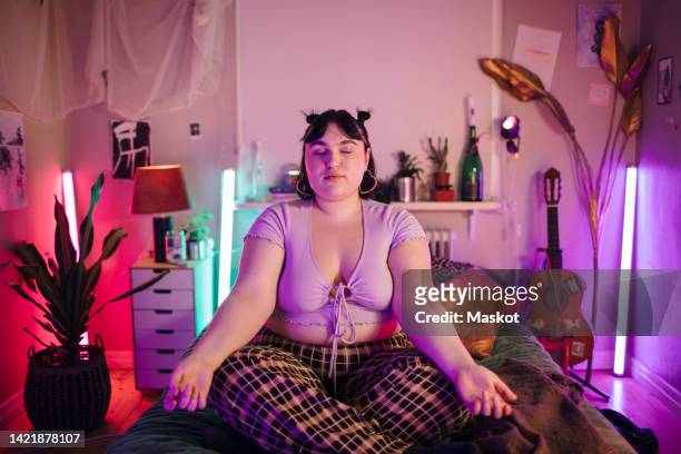 Young woman meditating on bed in illuminated bedroom at home