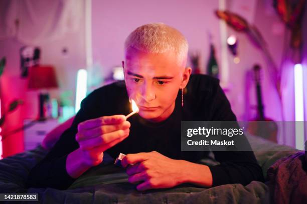 hipster young man looking at burning matchstick while lying on bed in illuminated bedroom - streichholzschachtel stock-fotos und bilder