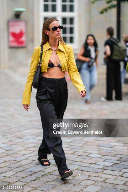 Guest wears black sunglasses from Balenciaga, a black pearls necklace, gold earrings, a yellow cropped shirt jacket, a black grained leather shoulder...