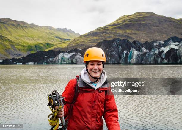 teenage boy wearing ice climbing gear smiling in front of a glacier lake and blue coloured ice glacier - boy in hard hat stock pictures, royalty-free photos & images