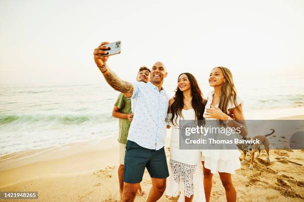 medium wide shot of smiling family taking selfie while standing on beach - funkwelle stock-fotos und bilder