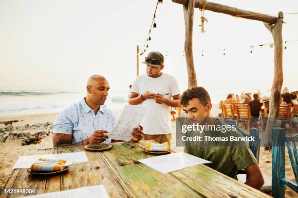 medium wide shot of waiter taking order from family at beach restaurant - family ordering at restaurant stock pictures, royalty-free photos & images