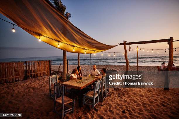 wide shot of smiling family at tropical beach restaurant at sunset - summer lights stockfoto's en -beelden