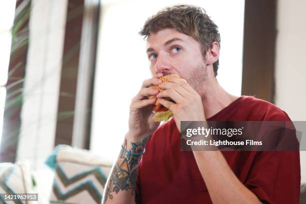 male with tattoos sits to eat savory burger on sesame bun - male burger eating fotografías e imágenes de stock