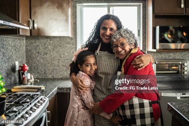 grandmother, mother and daughter embracing in the kitchen at home - hispanic grandmother stock pictures, royalty-free photos & images