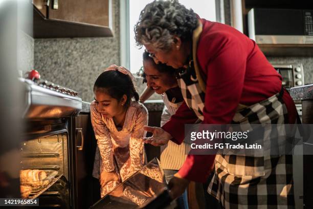 grandmother, mother and daughter looking at food in the oven at home - latin american culture stock pictures, royalty-free photos & images
