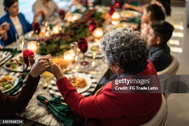 ältere frau mit familie, die an weihnachten zu hause am esstisch betet - religious service stock-fotos und bilder