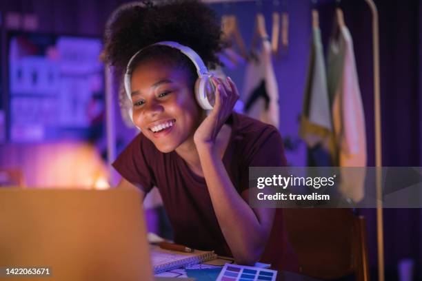 adolescente afro-americana usando um laptop e ouvindo streaming de música brincando com fones de ouvido navegando nas redes sociais em um escritório moderno à noite, momento de estilo de vida alegre à noite - upload - fotografias e filmes do acervo