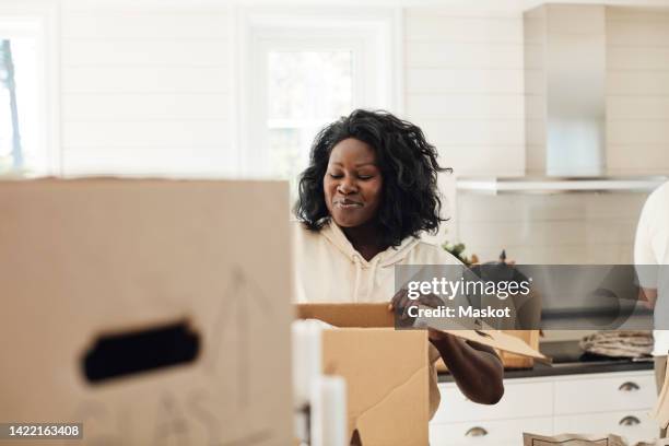smiling woman unpacking cardboard box while standing in kitchen at home - uitpakken activiteit stockfoto's en -beelden