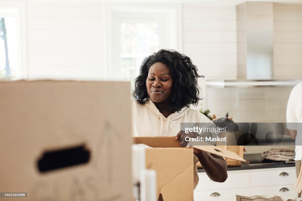 Smiling woman unpacking cardboard box while standing in kitchen at home