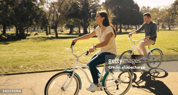 fit couple cycling on bicycles in a park, bonding and having a casual chat and free time outdoors. active husband and wife talking while exercising and doing cardio, a healthy lifestyle together - adult riding bike through park stock pictures, royalty-free photos & images
