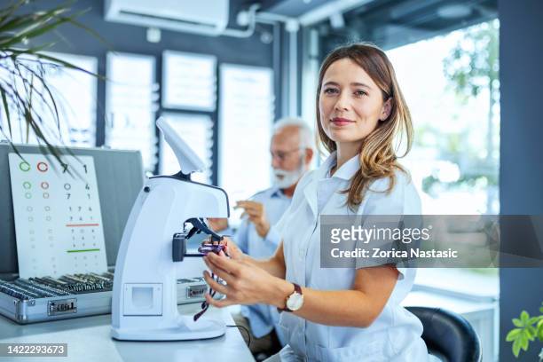 mujer joven midiendo vidrio en gafas para un paciente mayor mirando la cámara - optician fotografías e imágenes de stock