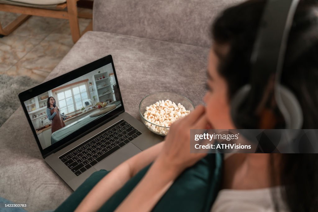 Over the shoulder view woman wearing headphone watching Movie Video on demand on laptop in the living room at home