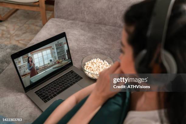 over the shoulder view woman wearing headphone watching movie video on demand on laptop in the living room at home - stream stock pictures, royalty-free photos & images