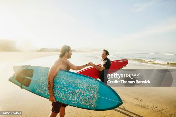 wide shot of smiling friends fist bumping after morning surf session - beach hold surfboard stock-fotos und bilder